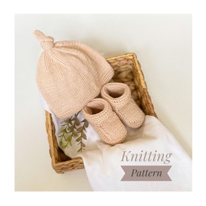 Peut inclure: Un ensemble de bonnet et chaussons en tricot pour bébé de couleur rose pâle. Le bonnet a un nœud sur le dessus et les chaussons ont un design simple. L'ensemble est présenté dans un panier en osier avec un tissu blanc et de la verdure.