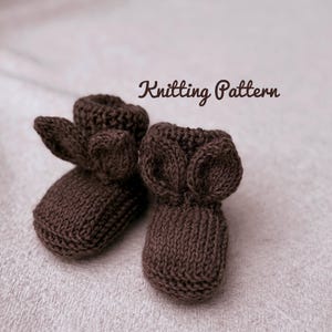 Könnte beinhalten: Ein Paar braune, gestrickte Babyschuhe mit ohrenförmigen Details. Der Text "Knitting Pattern" ist in einer Schreibschrift über den Schuhen abgebildet. Die Schuhe sind aus weichem, strukturiertem Garn gefertigt.