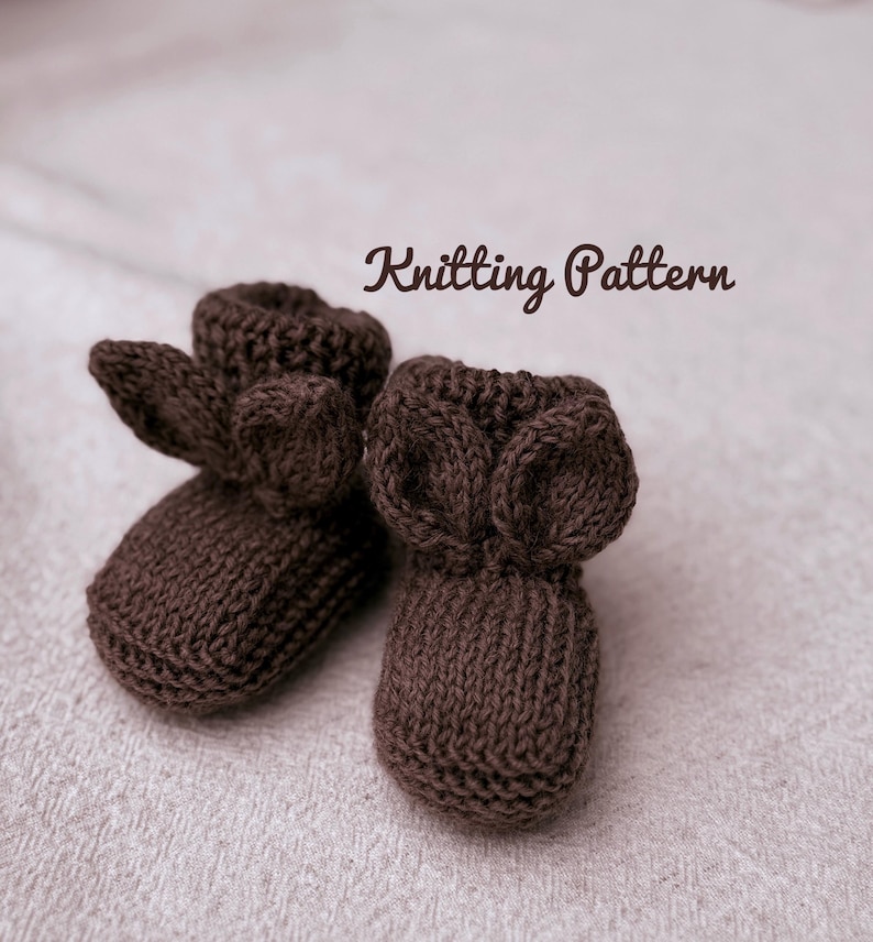 May include: A pair of brown knitted baby booties with ear-shaped accents. The text "Knitting Pattern" is displayed in a cursive font above the booties. The booties are made of a soft, textured yarn.
