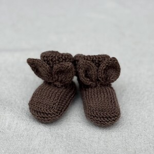 May include: A pair of brown knitted baby booties with ear-shaped accents. The booties are made of a soft, textured yarn and are designed to keep a baby's feet warm. The booties are shown on a light gray surface.