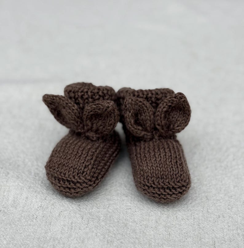 May include: A pair of brown knitted baby booties with ear-shaped accents. The booties are made of a soft, textured yarn and are designed to keep a baby's feet warm. The booties are shown on a light gray surface.