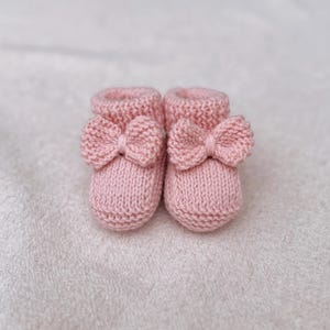 May include: A pair of soft pink knitted baby booties. Each bootie features a decorative bow on the front. The booties are made of a textured yarn and are designed for newborns. The background is a soft, white fabric.