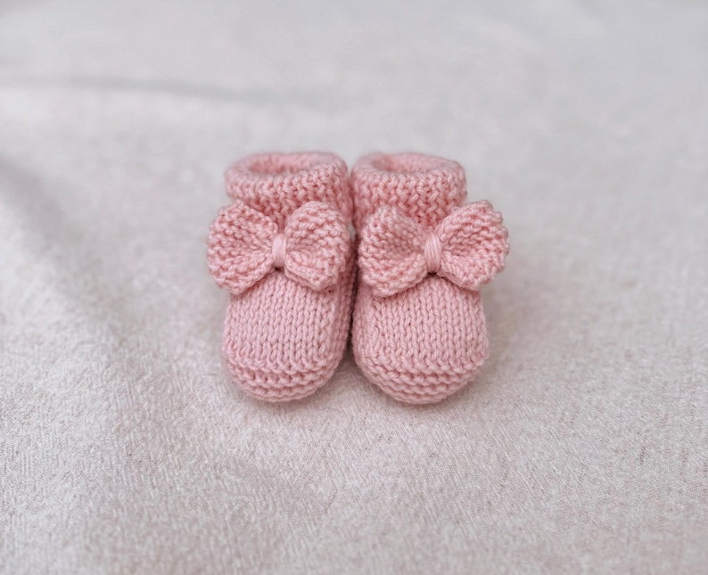 May include: A pair of soft pink knitted baby booties. Each bootie features a decorative bow on the front. The booties are made of a textured yarn and are designed for newborns. The background is a soft, white fabric.
