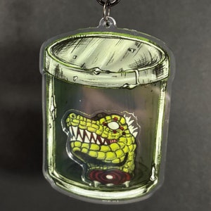 Könnte beinhalten: Ein durchsichtiger Acryl-Schlüsselanhänger mit einem grünen und gelben Cartoon-Dinosaurierkopf in einem Glasgefäß. Der Dinosaurier hat rote Augen und scharfe Zähne.