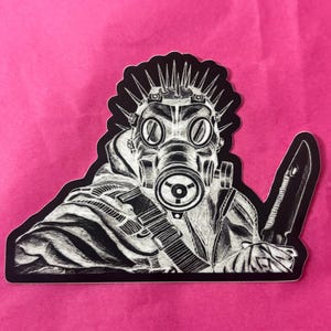 May include: Black and white sticker depicting a figure in a gas mask and spiked headgear, holding a large knife. The sticker has a black border and is set against a pink background. The illustration style is detailed and graphic.