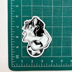 May include: A black and white sticker featuring a rat wearing a hat and goggles, perched atop a star-shaped design. The rat has a long tail and detailed claws. The sticker is on a green cutting mat.