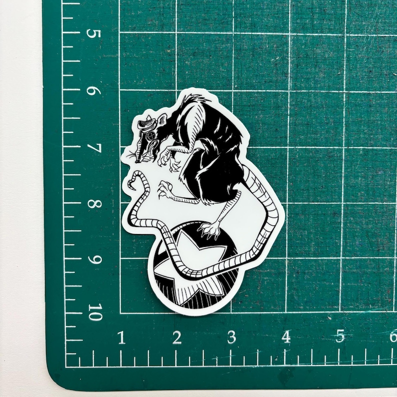 May include: A black and white sticker featuring a rat wearing a hat and goggles, perched atop a star-shaped design. The rat has a long tail and detailed claws. The sticker is on a green cutting mat.