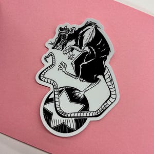 May include: Black and white sticker featuring a rat wearing a hat, with a star design. The rat is perched atop a star-shaped design. The sticker is on a pink background.