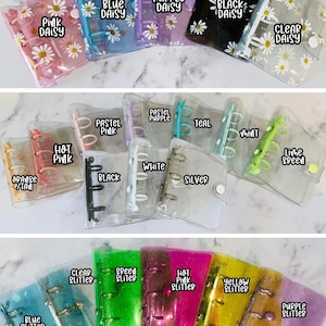 May include: A collection of mini binders in various colors, including pink, blue, purple, black, and clear. Each binder features a daisy pattern and is labeled with its color. Some binders have glitter.