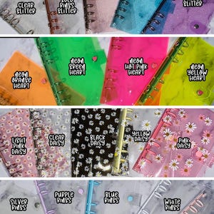 May include: A collection of clear plastic A6 disc bound notebooks with various designs. The designs include glitter, hearts, daisies, and different colored rings. The text on the covers includes "Clear Glitter", "Rose Gold Rings Glitter", "Pink Glitter", "Blue Glitter", "Purple Glitter", "Neon Orange Heart", "Neon Green Heart", "Neon Hot Pink Heart", "Neon Yellow Heart", "Light Pink Daisy", "Clear Daisy", "Black Daisy", "Yellow Daisy", "Pink Daisy", "Silver Rings", "Purple Rings", "Blue Rings", "White Rings", and "Pastel Pink Rings".