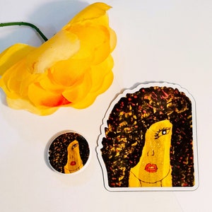 May include: A sticker and a button featuring a colorful illustration of a woman with a large afro. The woman has a yellow face with a red mouth and one blue eye. The background is black with red and yellow accents.