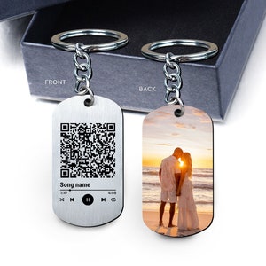 May include: A silver keychain with a black and white QR code on the front and a photo of a couple on the back. The front also has a music player interface with the text "Song name", "1:10", and "4:08".