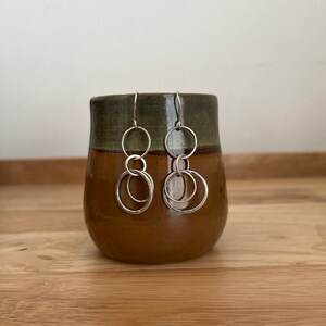 Puede incluir: Pendientes de plata con tres círculos interconectados. Los pendientes se muestran en una taza de cerámica marrón.