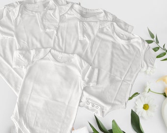 May include: Four white blank t-shirts, two with short sleeves and two with long sleeves, laid out on a white surface with green leaves and white flowers.