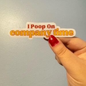 May include: A white sticker with the text "I Poop On company time" in orange and yellow lettering. The sticker is held by a hand with red nail polish against a light blue background.