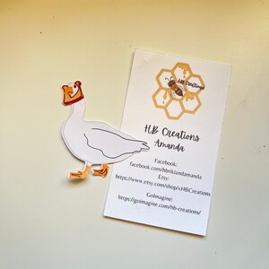 May include: A white goose with an orange beak and feet wearing a brown and orange hat. The goose is a sticker with a white background. The sticker is on a white card with the text "HB Creations Amanda" and contact information for Facebook, Etsy, and Goimagine.