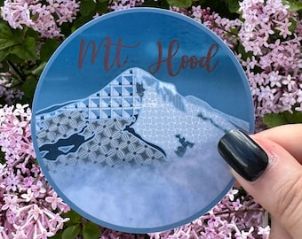 May include: A round sticker with a blue background and a white mountain with a geometric pattern. The text "Mt Hood" is written in brown letters on the sticker.