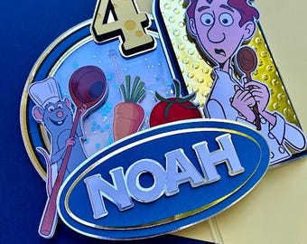 May include: A blue and gold cake topper featuring Remy the rat from Ratatouille, a chef holding a spoon, a number 4, a carrot, a tomato, and the name Noah.