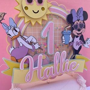 K&ouml;nnte beinhalten: Ein rosa und gelber Tortendekor mit einer Sonne, der Zahl eins, einem Minnie Mouse-Charakter mit Sonnenbrille, einem Daisy Duck-Charakter und dem Namen "Hallie".