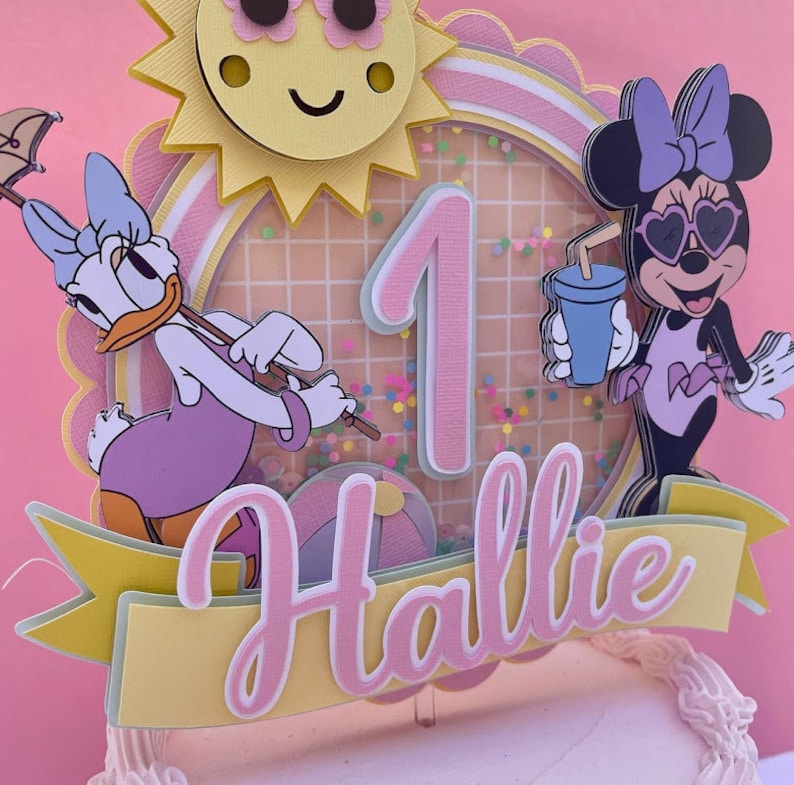 K&ouml;nnte beinhalten: Ein rosa und gelber Tortendekor mit einer Sonne, der Zahl eins, einem Minnie Mouse-Charakter mit Sonnenbrille, einem Daisy Duck-Charakter und dem Namen "Hallie".