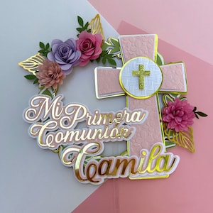 Pode incluir: Um topo de bolo rosa e dourado com uma cruz, flores e o texto "Mi Primera Comunión Camila".