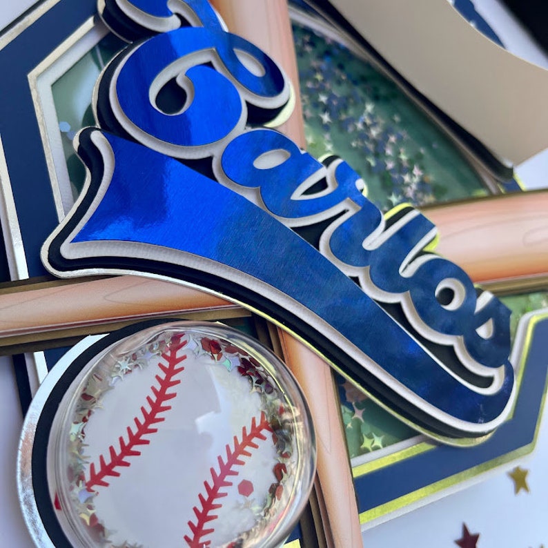 Pu&ograve; includere: Un biglietto a tema baseball blu e bianco con il nome "Carlos" in un font blu, bianco e nero. Il biglietto ha una palla da baseball con cuciture rosse e glitter all'interno di una cupola trasparente.