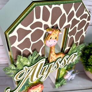 K&ouml;nnte beinhalten: Ein brauner und wei&szlig;er Giraffen-Druck-Kuchentopper mit gr&uuml;nem Blattwerk. Der Topper zeigt eine Giraffe, einen L&ouml;wen und ein Zebra, und der Name "Alyssa" ist in goldenen Buchstaben geschrieben.