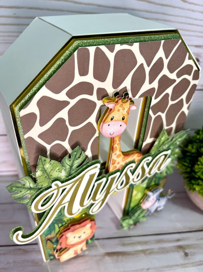 K&ouml;nnte beinhalten: Ein brauner und wei&szlig;er Giraffen-Druck-Kuchentopper mit gr&uuml;nem Blattwerk. Der Topper zeigt eine Giraffe, einen L&ouml;wen und ein Zebra, und der Name "Alyssa" ist in goldenen Buchstaben geschrieben.