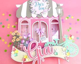 May include: A pink and gray papercraft cake topper featuring a small shop with a sign that reads "Par Tea" and a bicycle with flowers in the basket. The name "Gia" is written in pink on a yellow banner.