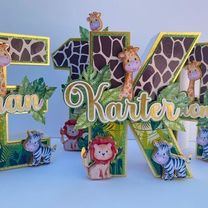 K&ouml;nnte beinhalten: Drei Holzbuchstaben, E, K und O, im Dschungel-Design. Die Buchstaben sind mit gr&uuml;nen Bl&auml;ttern, einer Giraffe, einem Zebra und einem L&ouml;wen verziert. Die Buchstaben ergeben den Namen "Ethan Karter Tony".