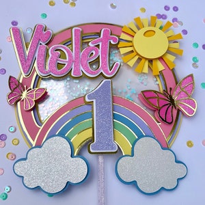 May include: A pink, purple and yellow rainbow cake topper with a glittery number one, the name "Violet" and a yellow sun with a pink butterfly on each side.