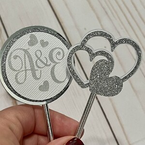 Puede incluir: Dos adornos para tartas plateados, uno con un diseño circular y las letras "A & E" y el otro con un diseño de doble corazón.