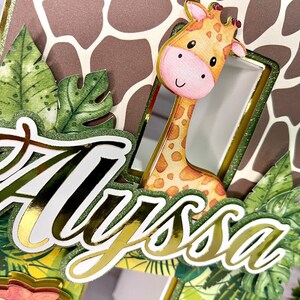 K&ouml;nnte beinhalten: Ein goldener Glitzer-Kuchentopper mit dem Namen "Alyssa" in einer kursiven Schrift. Der Topper zeigt eine Giraffe, die hinter gr&uuml;nen Bl&auml;ttern hervorlugt, und einen braunen und wei&szlig;en Giraffenmuster-Hintergrund.