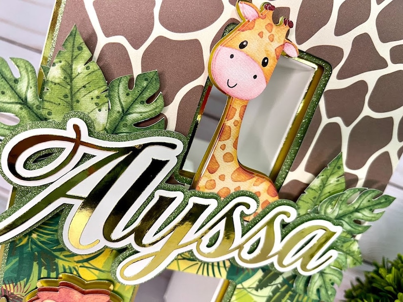 K&ouml;nnte beinhalten: Ein goldener Glitzer-Kuchentopper mit dem Namen "Alyssa" in einer kursiven Schrift. Der Topper zeigt eine Giraffe, die hinter gr&uuml;nen Bl&auml;ttern hervorlugt, und einen braunen und wei&szlig;en Giraffenmuster-Hintergrund.