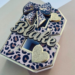 Puede incluir: Una caja de regalo decorativa con un diseño de estampado de leopardo en rosa, marrón y negro. La caja presenta un lazo, dos corazones dorados con purpurina y el nombre "Blake" en letras doradas con purpurina. La caja tiene un borde dorado y una ventana.