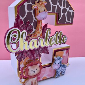 K&ouml;nnte beinhalten: Ein wei&szlig;er Papp-Kuchentopper in Form der Zahl Zwei, verziert mit einer Giraffe, einem L&ouml;wen und einem Zebra. Die Zwei ist mit einem rosa-braunen Giraffenmuster verziert. Der Name Charlotte ist in goldenen Buchstaben auf dem Kuchentopper geschrieben.