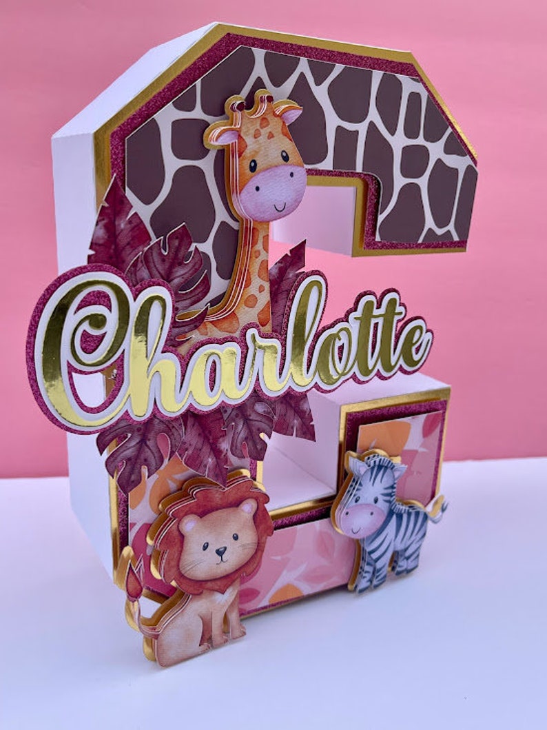 K&ouml;nnte beinhalten: Ein wei&szlig;er Papp-Kuchentopper in Form der Zahl Zwei, verziert mit einer Giraffe, einem L&ouml;wen und einem Zebra. Die Zwei ist mit einem rosa-braunen Giraffenmuster verziert. Der Name Charlotte ist in goldenen Buchstaben auf dem Kuchentopper geschrieben.