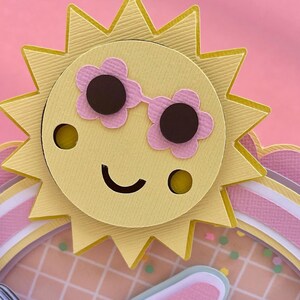 K&ouml;nnte beinhalten: Eine gelbe Papiersonne mit einem l&auml;chelnden Gesicht, die eine rosa Sonnenbrille tr&auml;gt. Die Sonne ist auf einen rosa-wei&szlig; gestreiften Papierhintergrund gelegt.