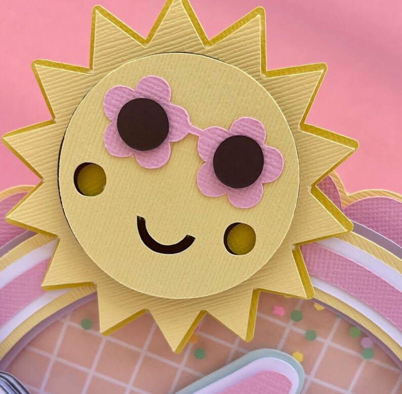 K&ouml;nnte beinhalten: Eine gelbe Papiersonne mit einem l&auml;chelnden Gesicht, die eine rosa Sonnenbrille tr&auml;gt. Die Sonne ist auf einen rosa-wei&szlig; gestreiften Papierhintergrund gelegt.