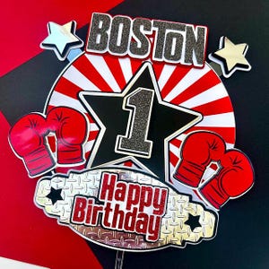 May include: A birthday cake topper with a boxing theme. The topper features the word "BOSTON" in black glitter, a large black star with the number "1", and red boxing gloves. The words "Happy Birthday" are also included.
