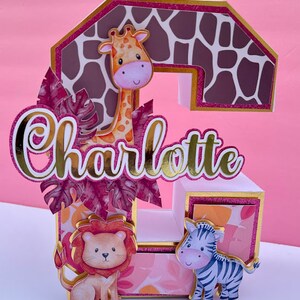 K&ouml;nnte beinhalten: Ein rosa-goldener 3D-Tortenaufsatz mit dem Namen "Charlotte" in goldenen Buchstaben. Der Aufsatz zeigt eine Giraffe, einen L&ouml;wen und ein Zebra, alle mit rosa und orangefarbenen Akzenten. Der Hintergrund ist ein rosa-oranger Giraffenprint.
