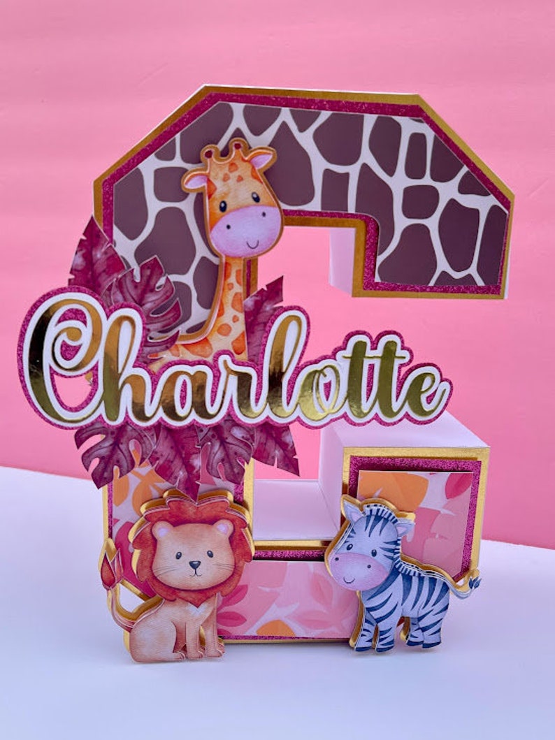 K&ouml;nnte beinhalten: Ein rosa-goldener 3D-Tortenaufsatz mit dem Namen "Charlotte" in goldenen Buchstaben. Der Aufsatz zeigt eine Giraffe, einen L&ouml;wen und ein Zebra, alle mit rosa und orangefarbenen Akzenten. Der Hintergrund ist ein rosa-oranger Giraffenprint.