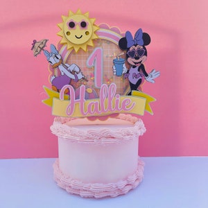 K&ouml;nnte beinhalten: Ein rosa und gelber Tortendekor mit einer l&auml;chelnden Sonne, der Zahl eins, einem Minnie Mouse-Charakter mit Sonnenbrille und Badeanzug und einer Entenfigur, die einen Regenschirm h&auml;lt. Der Topper tr&auml;gt den Schriftzug "Hallie" auf einem gelben Banner.