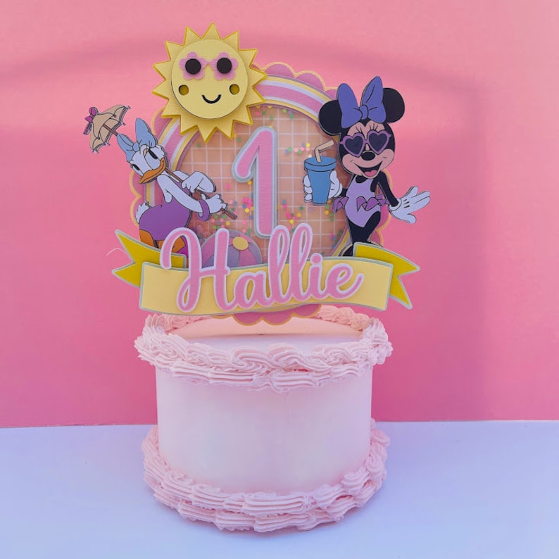 K&ouml;nnte beinhalten: Ein rosa und gelber Tortendekor mit einer l&auml;chelnden Sonne, der Zahl eins, einem Minnie Mouse-Charakter mit Sonnenbrille und Badeanzug und einer Entenfigur, die einen Regenschirm h&auml;lt. Der Topper tr&auml;gt den Schriftzug "Hallie" auf einem gelben Banner.