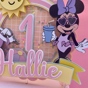 K&ouml;nnte beinhalten: Ein rosa und gelber Tortendekor mit einer Comicfigur, die eine Sonnenbrille und einen Badeanzug tr&auml;gt, der Zahl eins und dem Namen "Hallie".