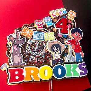 May include: A vibrant cake topper with characters from the film Coco, including Miguel, Dante, and Hector. The topper features the name "Brooks" in rainbow colours, the number "4", and the phrase "Un Poco Loco".