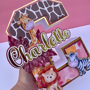 K&ouml;nnte beinhalten: Ein dreidimensionaler Buchstabe C in Pink, Braun und Gold mit Glitzerakzenten. Der Buchstabe ist mit einer Giraffe, einem L&ouml;wen und einem Zebra verziert. Der Name "Charlotte" ist in Goldglitter auf den Buchstaben geschrieben.