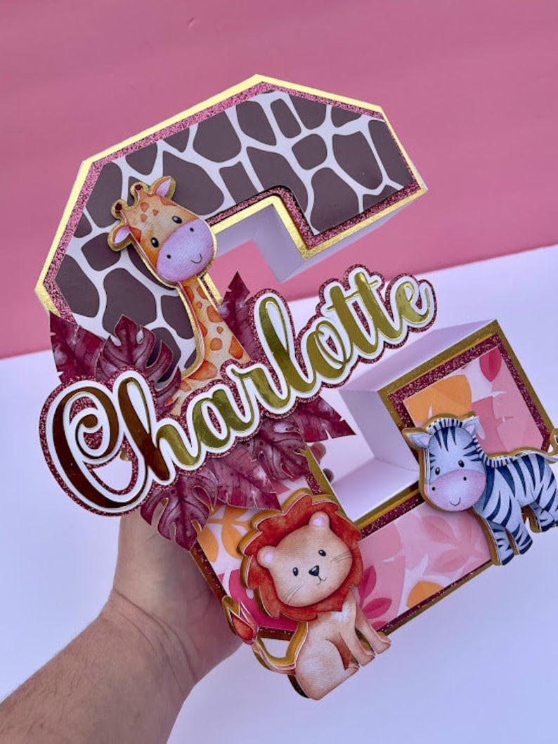 K&ouml;nnte beinhalten: Ein dreidimensionaler Buchstabe C in Pink, Braun und Gold mit Glitzerakzenten. Der Buchstabe ist mit einer Giraffe, einem L&ouml;wen und einem Zebra verziert. Der Name "Charlotte" ist in Goldglitter auf den Buchstaben geschrieben.