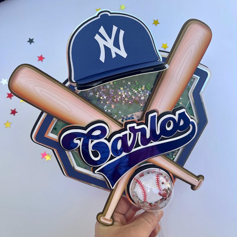Pu&ograve; includere: Un cappello da baseball blu e bianco con il logo dei New York Yankees, due mazze da baseball di legno, un campo da baseball blu e bianco e il nome Carlos in lettere blu e bianche. Le mazze da baseball sono incrociate l'una sull'altra e il cappello da baseball &egrave; sopra le mazze. C'&egrave; una piccola palla da baseball con cuciture rosse e bianche al centro del design.