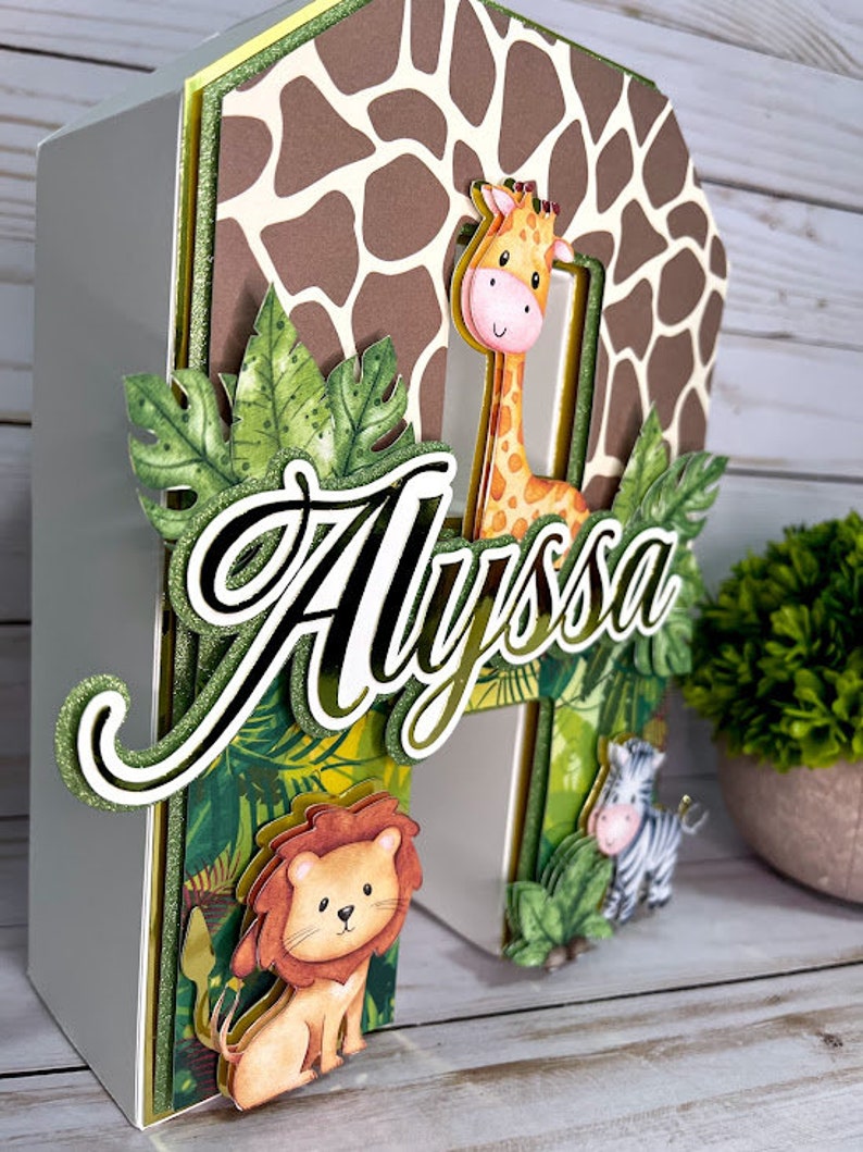 K&ouml;nnte beinhalten: Ein wei&szlig;er, dreidimensionaler Buchstabe "A", der mit einem Dschungel-Motiv verziert ist. Der Buchstabe ist mit gr&uuml;nen Bl&auml;ttern, einer Giraffe, einem L&ouml;wen und einem Zebra bedeckt. Der Name "Alyssa" ist in goldenen Glitzerbuchstaben auf der Vorderseite des Buchstabens geschrieben.